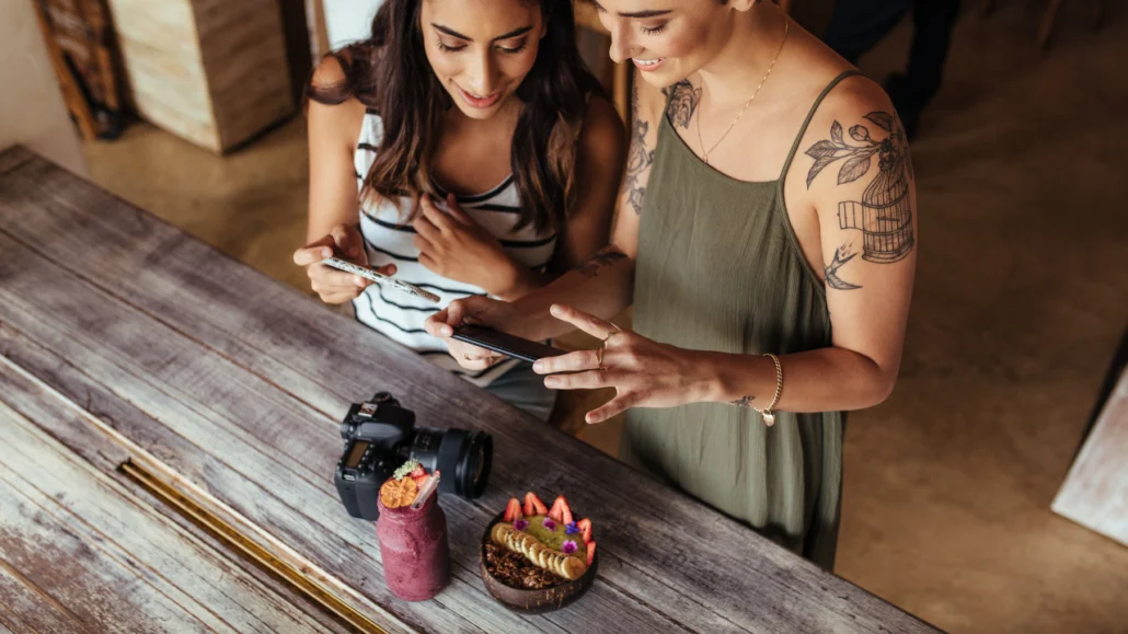 Photograph of two young women holding their smartphones and pointing them at a camera, a drink and a plate of food on a table. This image is a visual example of how User Generated Content is made, which is one of the basics for good local SEO.